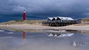 Roter Leuchtturm auf einer Düne mit einem modernen Strandpavillon im Vordergrund, dramatischer dunkler Himmel und Spiegelung im nassen Sand.